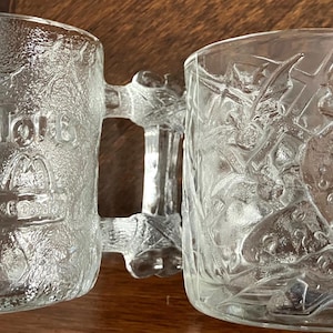 May include: Two clear glass mugs with textured surfaces. The left mug features the embossed text The Flintstones and a McDonald's logo. The right mug displays an embossed character wearing a hat. Both mugs have handles shaped like bones.