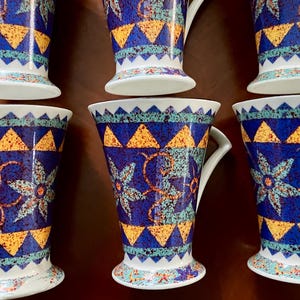 May include: Set of six ceramic mugs with a vibrant, geometric design. The mugs feature a deep blue background adorned with yellow triangles, teal accents, and floral patterns. Each mug has a white handle and base, creating a striking contrast.