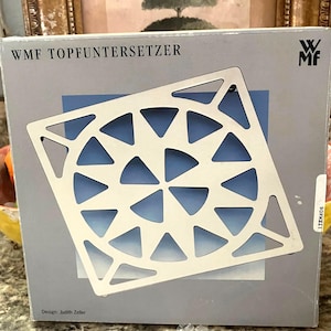May include: A silver metal trivet with a geometric design of triangles. The trivet is square shaped and has a WMF logo in the top right corner. The text "WMF TOPFUNTERSETZER" is printed at the top of the image. The text "Design: Judith Zeller" is printed at the bottom of the image.