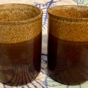 May include: Two brown and white ceramic mugs with a speckled glaze. The mugs have a wide, rounded shape and a handle.