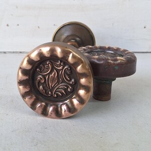 May include: Three antique copper-toned door knobs with ornate floral designs. The knobs have a round shape with detailed patterns and a warm, aged appearance. The background is a white, distressed door.
