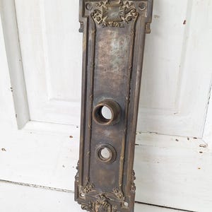 May include: Antique bronze door plate with ornate detailing. The rectangular plate features a lion's head at the top and decorative scrollwork. It has two circular holes for the door handle and lock. The finish is aged and shows signs of wear.