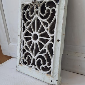 May include: An antique white cast iron vent cover with a decorative floral and geometric pattern. The rectangular frame has a distressed finish, revealing the dark metal beneath. A vintage home decor item.