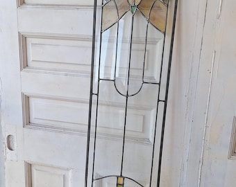 May include: A stained glass window panel with a geometric design featuring a central flower motif in shades of beige, green, and yellow. The panel is framed in black metal and is leaning against a white painted door.