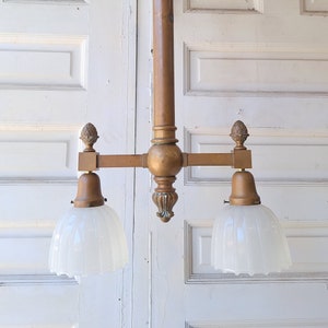 May include: A brass two-light chandelier with frosted glass shades. The chandelier has a long, vertical stem and a crossbar with two lights. The shades are bell-shaped and have a ribbed texture.