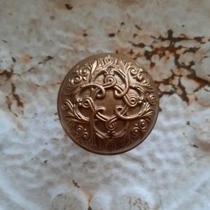 May include: A gold-toned metal knob with an intricate floral design. The knob is round and has a raised, ornate pattern.