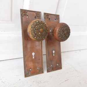May include: Two antique brass door knobs with ornate designs and keyholes. The knobs are attached to rectangular plates with decorative edges.