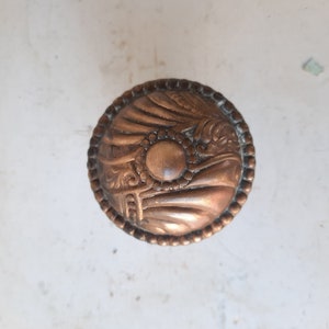 May include: A round, ornate, copper door knob with a raised, circular design in the center.