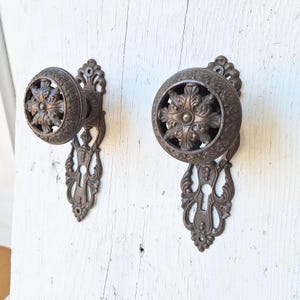 May include: Two antique bronze door knobs with ornate floral designs. The knobs are round with a detailed pattern, and the backplates have a keyhole and decorative flourishes. The hardware is mounted on a white, weathered wooden surface.