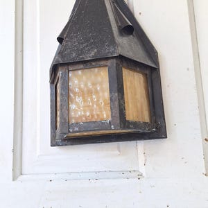 May include: A wall-mounted lantern featuring a dark, weathered metal frame with a pointed roof and textured, amber-colored glass panels. The fixture has a hexagonal shape and exhibits signs of age and oxidation on the metal surfaces.