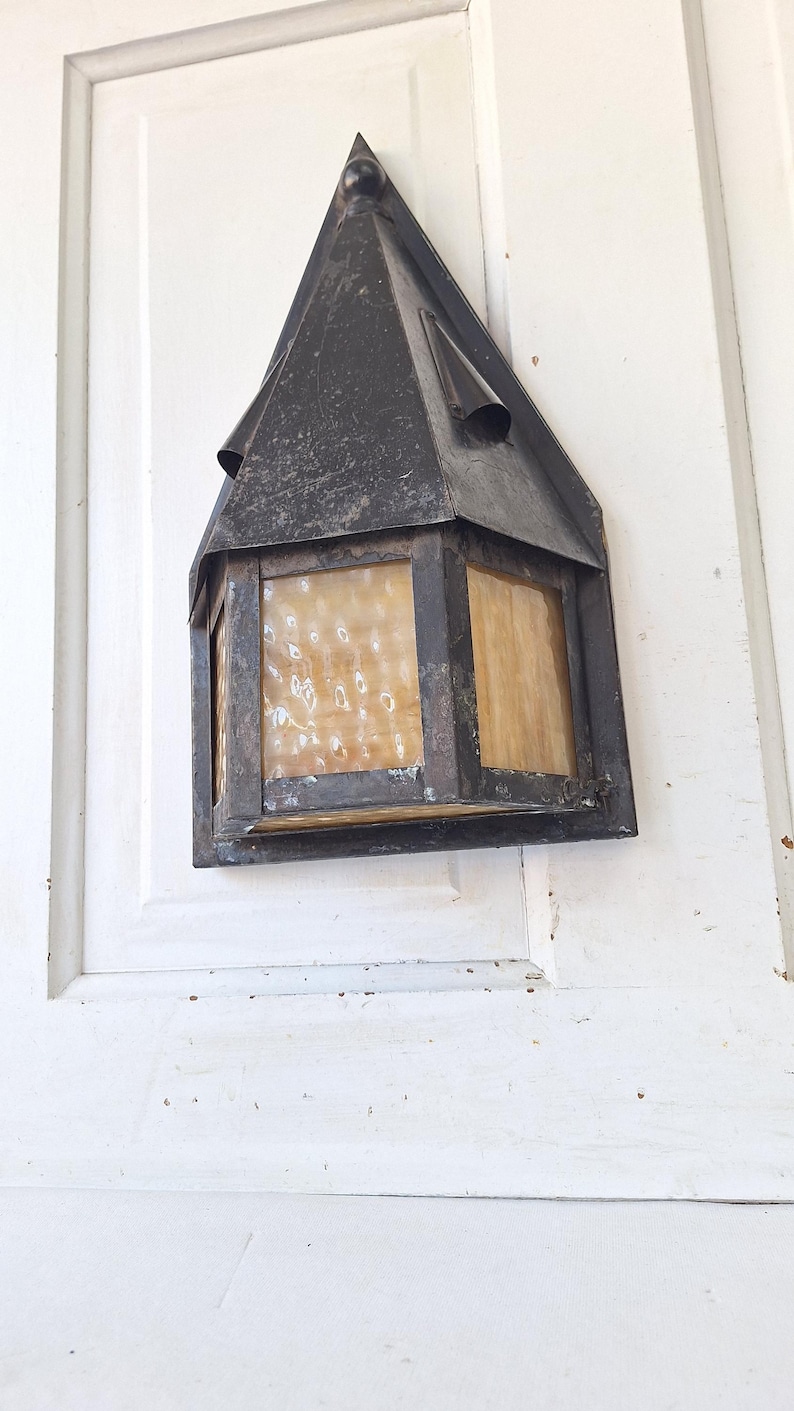 May include: A wall-mounted lantern featuring a dark, weathered metal frame with a pointed roof and textured, amber-colored glass panels. The fixture has a hexagonal shape and exhibits signs of age and oxidation on the metal surfaces.