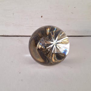 May include: A clear glass paperweight with a starburst design on the bottom. The paperweight has a round shape and a swirling, smoky interior. The glass reflects light, creating a play of colors.