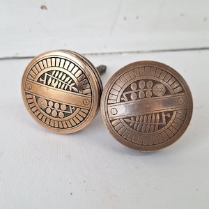 May include: Two antique bronze-colored cabinet knobs with intricate geometric designs. Each knob is round with a detailed pattern of lines, circles, and a central horizontal band. The knobs are likely made of metal and are suitable for furniture restoration.
