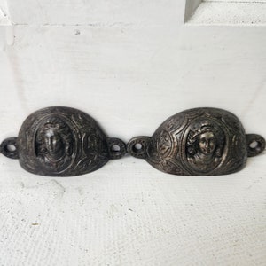 May include: Two antique metal drawer pulls with ornate designs featuring a woman's face in the center. The pulls are a dark bronze color and have a slightly curved shape.