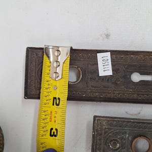 May include: Antique bronze door hardware, including a rectangular plate with a keyhole and decorative detailing. The plate is approximately 3 inches long. A yellow measuring tape is placed on the plate. The hardware has a vintage aesthetic.