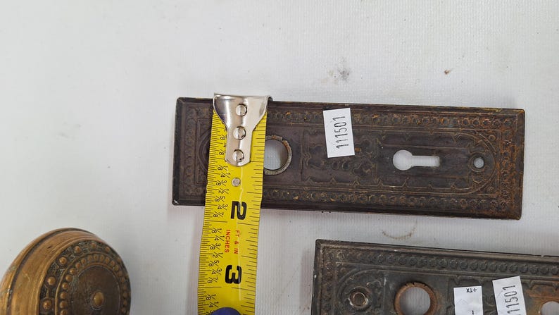 May include: Antique bronze door hardware, including a rectangular plate with a keyhole and decorative detailing. The plate is approximately 3 inches long. A yellow measuring tape is placed on the plate. The hardware has a vintage aesthetic.