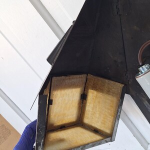 May include: A black metal wall-mounted lantern fixture featuring a triangular frame and translucent amber-colored panels. The structure includes a central light socket with brown wiring and a pointed top finial, showcasing a weathered finish.