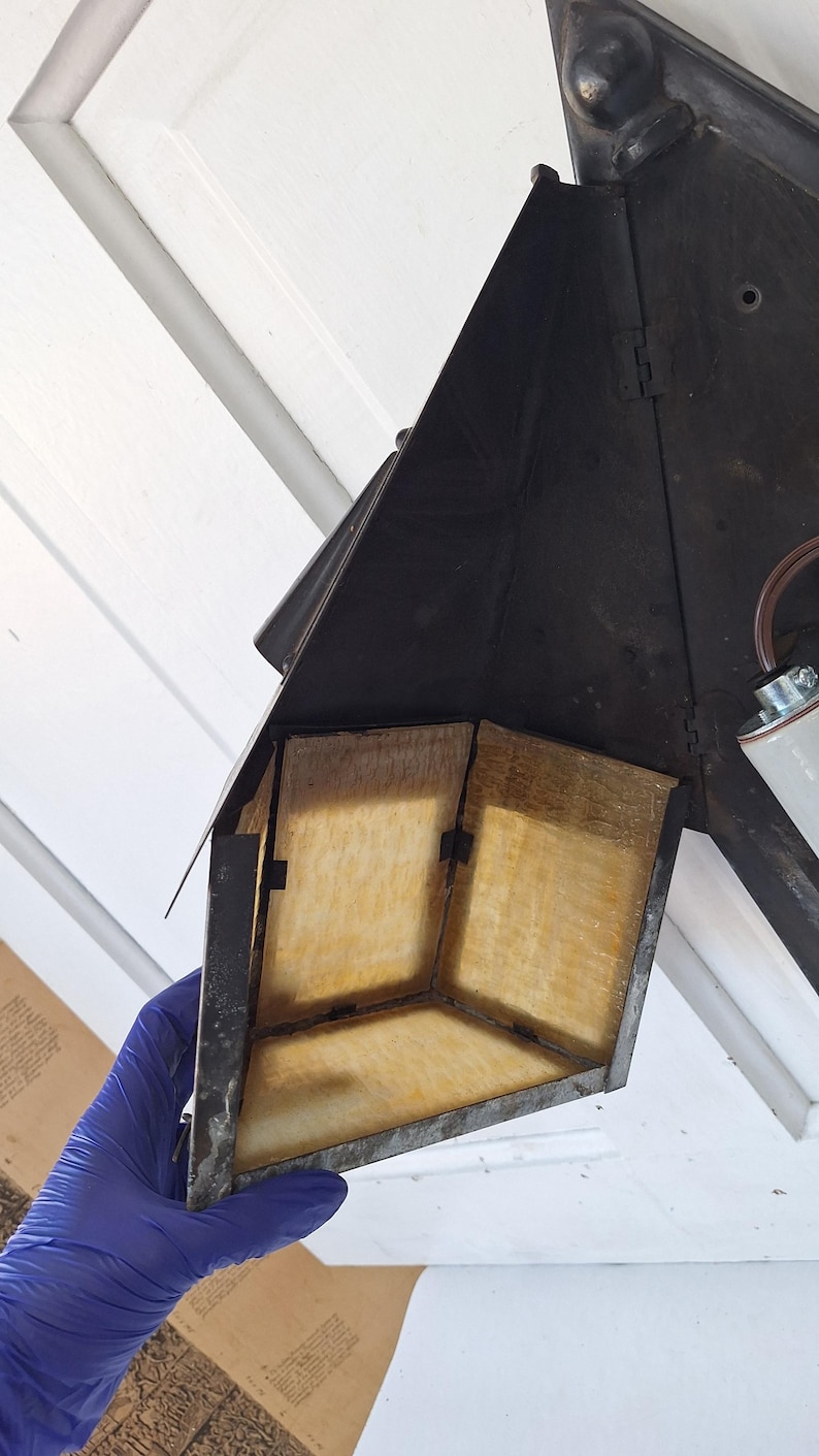 May include: A black metal wall-mounted lantern fixture featuring a triangular frame and translucent amber-colored panels. The structure includes a central light socket with brown wiring and a pointed top finial, showcasing a weathered finish.