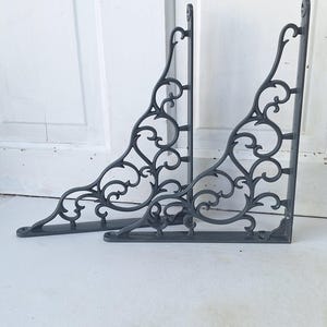 May include: Two dark gray, ornate metal shelf brackets. Each bracket features a decorative, swirling design. The brackets are designed to be mounted on a wall and provide support for shelves. The brackets are made of metal and have a matte finish.