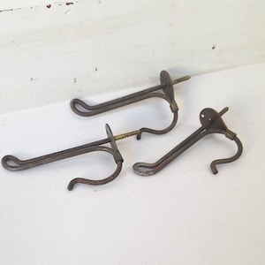 May include: Three dark brown metal wall hooks with a weathered finish. Each hook features a long, slender arm with a looped end and a circular mounting plate attached to a threaded screw for installation into a wall surface.