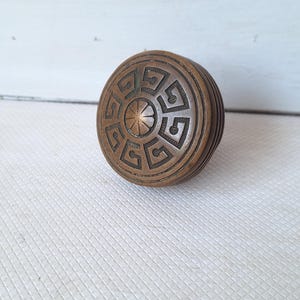 May include: A round, antique bronze door knob with a detailed geometric design. The knob has a circular pattern with a central starburst design. The sides of the knob are ribbed. The knob is likely made of metal.