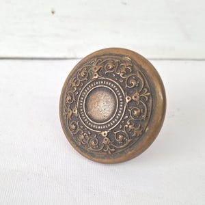 May include: Antique bronze-colored door knob with an ornate, circular design. The knob features a central circle surrounded by detailed floral and scrollwork patterns. The aged finish gives it a vintage aesthetic, suitable for restoration projects or adding a classic touch to any door.