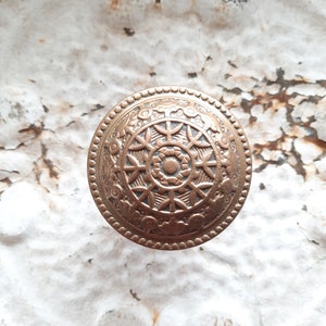 May include: A circular, gold-toned metal button with an intricate, floral design. The button has a raised center and a beaded edge.