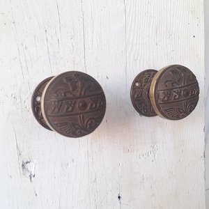 May include: Two round, dark bronze-toned metal door knobs featuring an intricate, embossed floral and geometric pattern. Each knob has a circular base plate with visible screw holes, showcasing a weathered, metallic finish with dark recessed details.