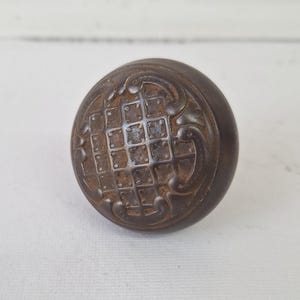 May include: A round, antique bronze door knob with a detailed, textured surface. The knob features a grid pattern in the center, surrounded by ornate scrollwork. The aged finish gives it a vintage aesthetic.