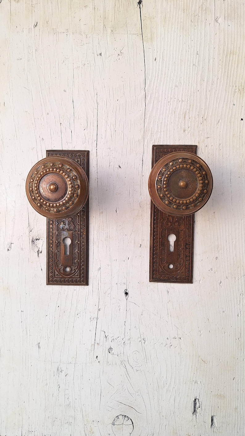 May include: Two antique bronze door knobs with rectangular backplates. The knobs are round with a central design surrounded by a beaded pattern. The backplates feature ornate detailing and keyholes. The door hardware is mounted on a white, weathered wooden surface.