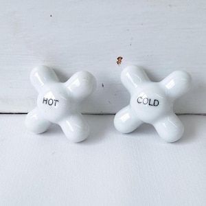 May include: Two white ceramic faucet handles with a cross-shaped design. One handle is labeled "HOT" in black text, and the other is labeled "COLD". The handles are set against a white background.