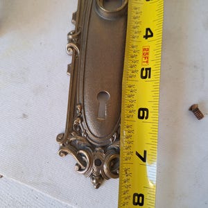 Puede incluir: Placa de puerta antigua de lat&oacute;n ornamentada con cerradura y adornos de volutas. La placa de la puerta mide aproximadamente 20 cm de largo. Se ve un tornillo cerca de la placa.