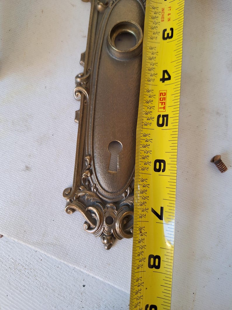 Puede incluir: Placa de puerta antigua de lat&oacute;n ornamentada con cerradura y adornos de volutas. La placa de la puerta mide aproximadamente 20 cm de largo. Se ve un tornillo cerca de la placa.