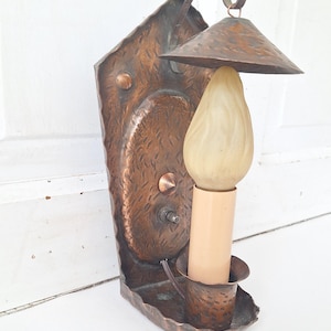 May include: A vintage copper wall sconce with a textured finish. The sconce features a decorative backplate, a small shade, and a light bulb. The fixture is mounted on a white surface.