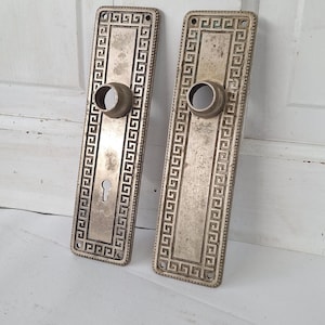 May include: Two antique bronze door handle backplates. Each rectangular plate features a keyhole, a circular handle opening, and a Greek key border design. The metal has a weathered, aged appearance.