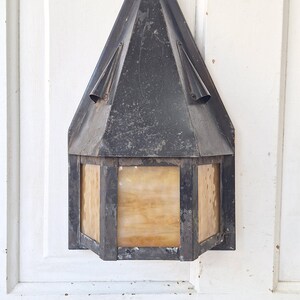 May include: A wall-mounted lantern featuring a dark, weathered metal frame with a pointed, triangular roof and a decorative finial at the top. The fixture includes rectangular panels of translucent, amber-toned glass with a mottled texture.