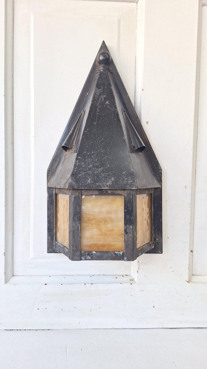 May include: A wall-mounted lantern featuring a dark, weathered metal frame with a pointed, triangular roof and a decorative finial at the top. The fixture includes rectangular panels of translucent, amber-toned glass with a mottled texture.