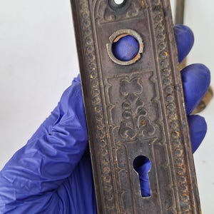 May include: An antique, rectangular door plate with ornate floral and geometric patterns. The metal plate is a dark, aged brown with signs of rust and wear. It features a keyhole and two screw holes, suggesting its use on a door.