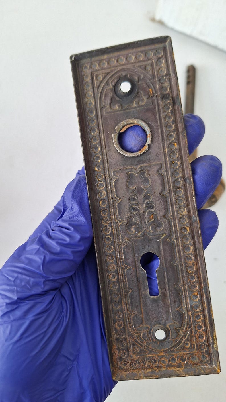 May include: An antique, rectangular door plate with ornate floral and geometric patterns. The metal plate is a dark, aged brown with signs of rust and wear. It features a keyhole and two screw holes, suggesting its use on a door.