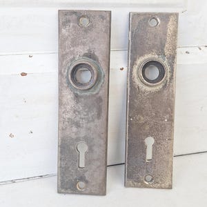 May include: Two antique, rectangular door plates with keyholes and circular openings for the doorknob. The metal plates have a weathered, aged appearance with a mottled, gray-brown patina, suggesting they are vintage or reclaimed architectural hardware.