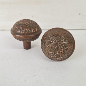 May include: Two antique bronze door knobs with intricate floral and geometric patterns. One knob is upright, showing the stem, while the other lies on its side. The knobs have a weathered, aged appearance, suggesting they are vintage.