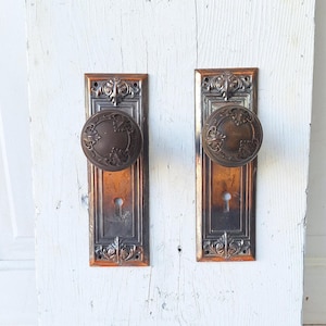 May include: A pair of antique door knobs with ornate details. The knobs are made of metal and have a silver finish. The backplates are rectangular and have a copper finish.