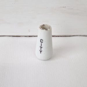 May include: A white, ceramic, cone-shaped object with the word "CITY" printed vertically in black. The top rim has a speckled, dark pattern. The object is set against a white background.