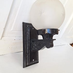 May include: An antique black wall sconce with a frosted glass globe. The globe has starburst designs. The sconce has a decorative bracket and mounting plate. A vintage lighting fixture.