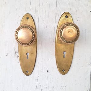 May include: Two antique brass door knobs with oval backplates. The knobs are round with a decorative beaded edge. The backplates have a keyhole and are mounted on a white, weathered wooden surface. The hardware has a vintage, aged appearance.