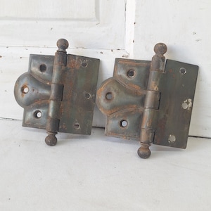 May include: Two antique metal hinges with a distressed, aged appearance. The hinges are a dark gray color with visible rust, featuring a decorative design and ball finials. Ideal for vintage decor or restoration projects.