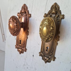 Puede incluir: Dos manijas de puerta antiguas con detalles ornamentados. Una es de color cobre, la otra de lat&oacute;n. Ambas tienen cerraduras y est&aacute;n montadas sobre una superficie de madera blanca desgastada. Las manijas tienen forma ovalada con patrones decorativos.