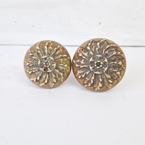 May include: Two antique brass door knobs with a floral design. The knobs are round and have a detailed, embossed pattern of leaves and petals. The brass has a warm, aged patina, suggesting vintage or retro style.
