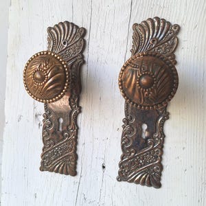 May include: Two matching antique-style door knobs with rectangular backplates, crafted from a dark bronze-toned metal. Each piece features intricate embossed floral and fan-shaped patterns, a central circular knob, and a keyhole opening.