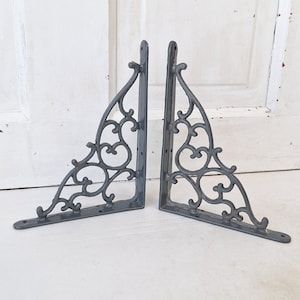 May include: Two gray, ornate metal shelf brackets. Each bracket has a triangular shape with decorative scrollwork and a flat base with pre-drilled holes for mounting. The brackets are designed for supporting shelves and adding a vintage aesthetic.